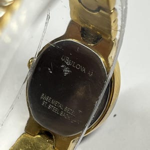 Może przedstawiać: Złoty zegarek Bulova z okrągłą tarczą i złotą bransoletą. Tarcza zegarka jest ciemna z widoczną nazwą marki i innym tekstem. Ramka jest z metalu podstawowego, a tył ze stali nierdzewnej.