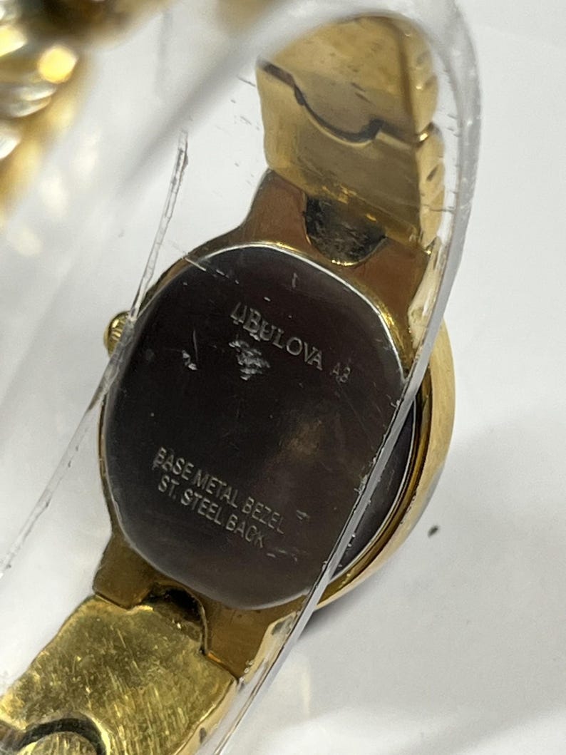 Może przedstawiać: Złoty zegarek Bulova z okrągłą tarczą i złotą bransoletą. Tarcza zegarka jest ciemna z widoczną nazwą marki i innym tekstem. Ramka jest z metalu podstawowego, a tył ze stali nierdzewnej.