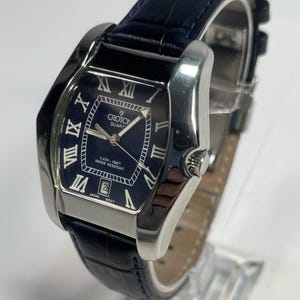 May include: A Croton Quartz watch with a rectangular navy blue face and silver case. It has Roman numeral hour markers, a date display, and a navy blue leather strap. Water-resistant to 5 ATM, equivalent to 150 feet.