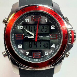 Peut inclure: Une montre num&eacute;rique argent&eacute;e avec un bracelet noir et une lunette rouge. Le cadran affiche l'heure et d'autres fonctions. La montre poss&egrave;de plusieurs boutons et une &eacute;chelle tachym&eacute;trique. La montre est marqu&eacute;e "JAPAN".