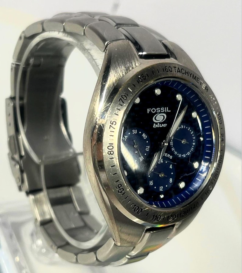 May include: A stainless steel Fossil Blue watch with a dark blue face. The watch features a tachymeter bezel and multiple subdials. The watch band is made of stainless steel.