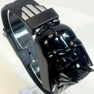 Pu&ograve; includere: Un orologio da polso nero con un design a casco di Darth Vader. L'orologio ha un cinturino nero con un motivo strutturato e una chiusura color argento. Il casco di Darth Vader &egrave; il punto focale, con dettagli intricati.