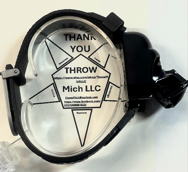 Pu&ograve; includere: Un porta orologio nero con una faccia trasparente, con il testo "THANK YOU" e "THROW Mich LLC". Il supporto ha un design di Darth Vader sul lato. Il porta orologio &egrave; progettato per contenere un orologio.