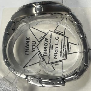 May include: A silver-toned wristwatch with a metal band, displayed in a clear plastic case. The watch face is obscured by a printed insert with the text "THANK YOU" and "THROW Mich LLC". The watch is new.