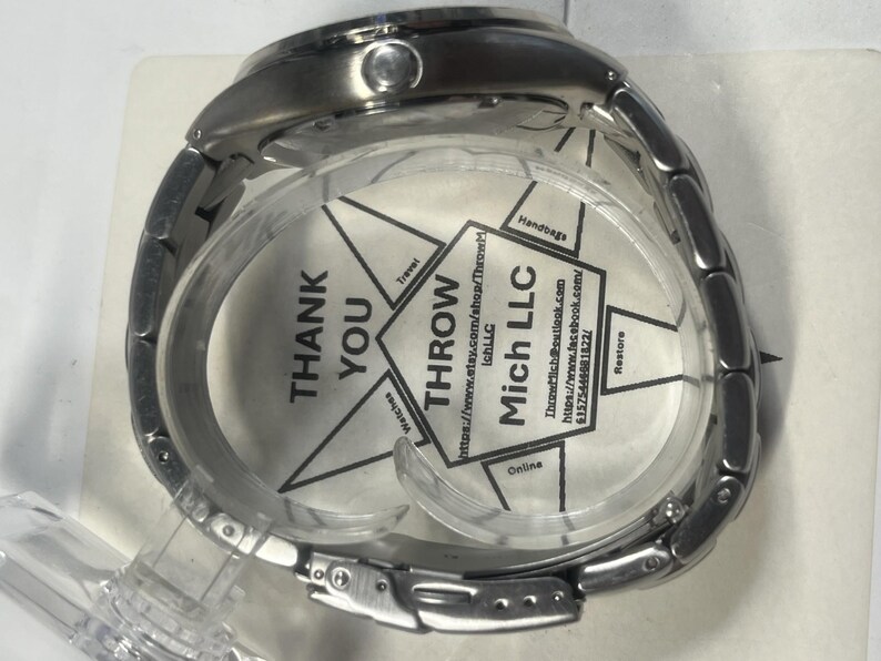 May include: A silver-toned wristwatch with a metal band, displayed in a clear plastic case. The watch face is obscured by a printed insert with the text "THANK YOU" and "THROW Mich LLC". The watch is new.