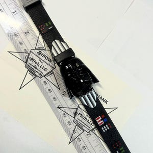 Pu&ograve; includere: Un orologio nero di Star Wars Darth Vader con un cinturino strutturato. L'orologio presenta una testa di Darth Vader e dettagli argentati. Un righello &egrave; posto accanto all'orologio, con il testo "THROW" e "Mich LLC" visibile.
