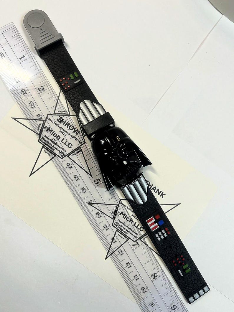 Pu&ograve; includere: Un orologio nero di Star Wars Darth Vader con un cinturino strutturato. L'orologio presenta una testa di Darth Vader e dettagli argentati. Un righello &egrave; posto accanto all'orologio, con il testo "THROW" e "Mich LLC" visibile.