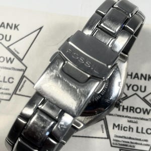 May include: A silver-toned Fossil wristwatch with a metal band. The watch is viewed from the back, showing the clasp and the brand name. The watch is on a white surface with some text in the background.
