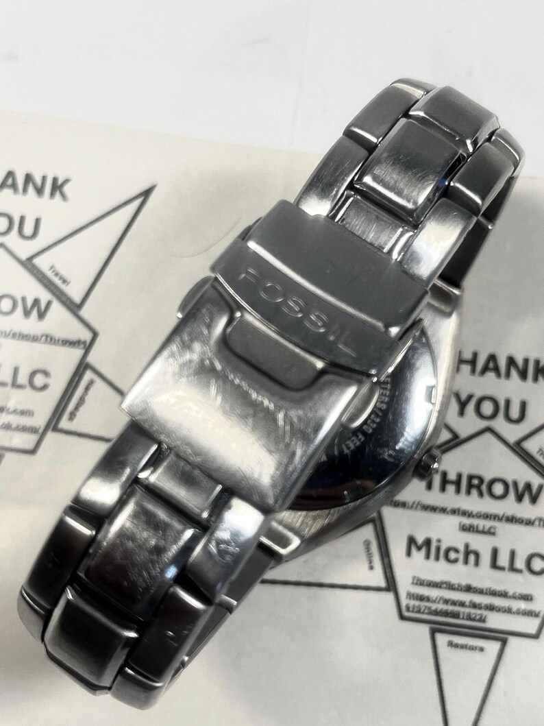 May include: A silver-toned Fossil wristwatch with a metal band. The watch is viewed from the back, showing the clasp and the brand name. The watch is on a white surface with some text in the background.