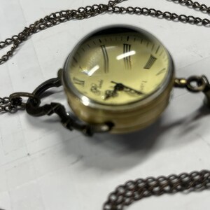 May include: A vintage-style pocket watch pendant necklace. The watch has a round, gold-toned case with a cream-colored face and Roman numeral hour markers. The watch is suspended from a long, dark chain.