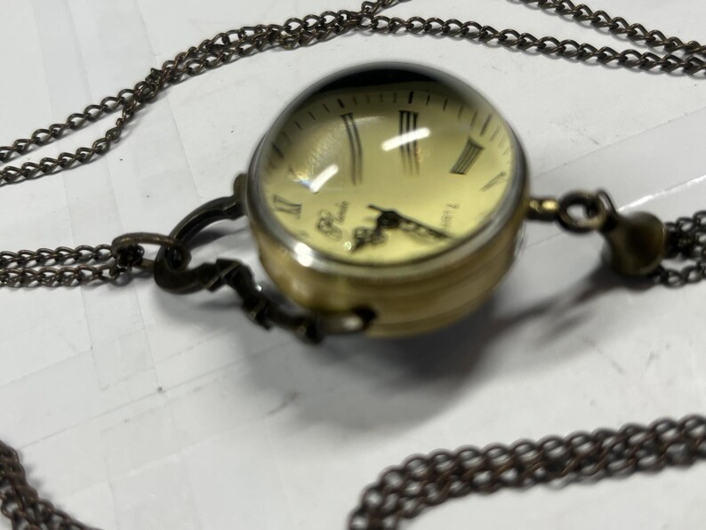 May include: A vintage-style pocket watch pendant necklace. The watch has a round, gold-toned case with a cream-colored face and Roman numeral hour markers. The watch is suspended from a long, dark chain.
