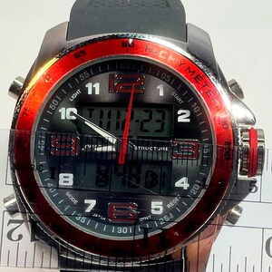 Peut inclure: Une montre-bracelet noire et argent&eacute;e avec une lunette rouge. Le cadran affiche l'heure num&eacute;rique et analogique, avec le texte "TACHYMETER" et "STRUCTURE". La montre a un bracelet en caoutchouc noir et plusieurs boutons.