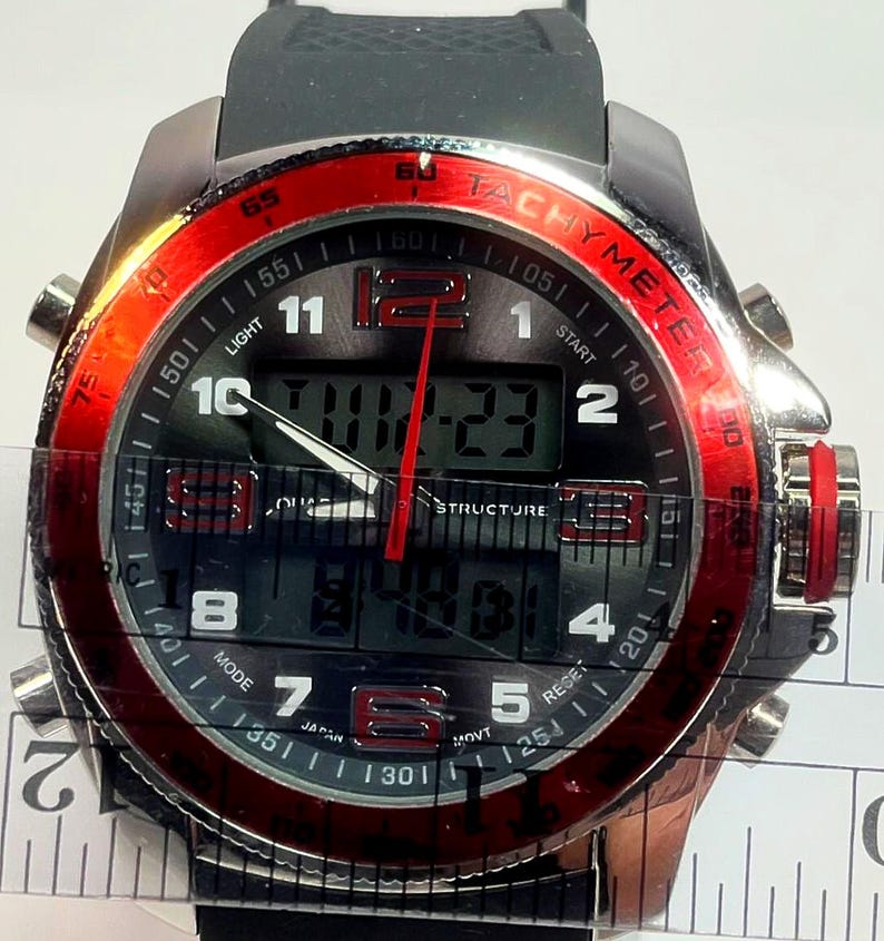 Peut inclure: Une montre-bracelet noire et argent&eacute;e avec une lunette rouge. Le cadran affiche l'heure num&eacute;rique et analogique, avec le texte "TACHYMETER" et "STRUCTURE". La montre a un bracelet en caoutchouc noir et plusieurs boutons.