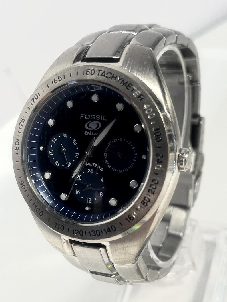 May include: A stainless steel Fossil watch with a dark blue face. The watch features a tachymeter bezel and multiple sub-dials. The watch face displays the brand name "Fossil" and the word "blue". The watch band is also stainless steel.