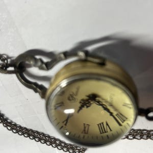 May include: A vintage-style pocket watch with a clear, domed face and Roman numeral hour markers. The watch has a gold-toned casing and a decorative chain. The watch face is cream-colored with black hands.