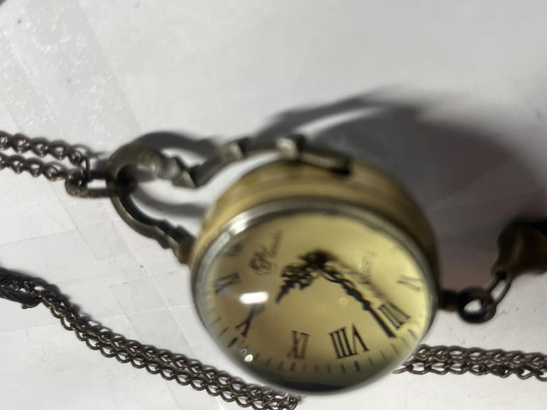 May include: A vintage-style pocket watch with a clear, domed face and Roman numeral hour markers. The watch has a gold-toned casing and a decorative chain. The watch face is cream-colored with black hands.