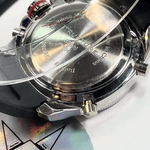 Peut inclure: Gros plan d'une montre en acier inoxydable avec un bracelet en caoutchouc noir. Le cadran affiche le texte "STRUQTURE" et "Made in China". La montre a plusieurs boutons et un accent rouge.