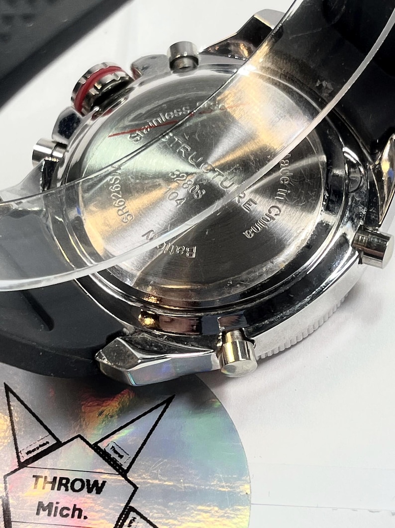 Peut inclure: Gros plan d'une montre en acier inoxydable avec un bracelet en caoutchouc noir. Le cadran affiche le texte "STRUQTURE" et "Made in China". La montre a plusieurs boutons et un accent rouge.