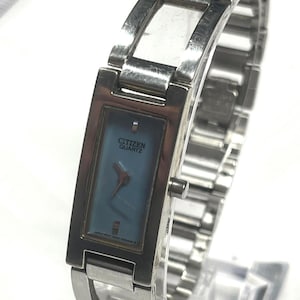May include: A silver Citizen Quartz wristwatch with a rectangular face and a light blue dial. The watch has a silver band and the brand name is visible on the watch face.