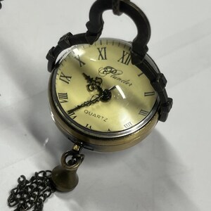 May include: A vintage-style quartz pocket watch with a cream-colored face and Roman numerals. The watch has a bronze-colored metal casing and a decorative chain with a tassel. The word "Plunder" is printed on the watch face.