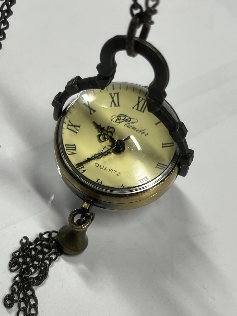 May include: A vintage-style quartz pocket watch with a cream-colored face and Roman numerals. The watch has a bronze-colored metal casing and a decorative chain with a tassel. The word "Plunder" is printed on the watch face.
