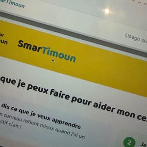 Puede incluir: Una pantalla de ordenador muestra el texto "SmarTimoun" en una pancarta amarilla. El texto está en una fuente sans-serif con "Smar" en negro y "Timoun" en verde azulado. Otro texto en negro es visible debajo de la pancarta.