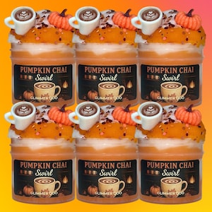 以下が含まれることがあります： 「Pumpkin Chai Swirl Glimmer Goo」とラベルが貼られたオレンジ色のスライムが入った6つの透明なプラスチックジャー。各ジャーの上部には、ラテアートが施された白いミニチュアのコーヒーカップと、オレンジ色のスライムの上に小さなオレンジ色のカボチャのチャームが飾られています。