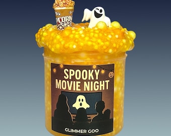 Könnte beinhalten: Ein durchsichtiger Kunststoffbehälter gefüllt mit gelbem, texturiertem Schleim, gekrönt von einem Miniatur-Popcorn-Eimer und einer weißen Geisterfigur. Das Etikett zeigt den Text SPOOKY MOVIE NIGHT, eine Geistergrafik auf einem Bildschirm und GLIMMER GOO.