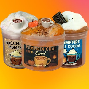 May include: Three clear plastic jars of textured slime labeled Macchiato Moment, Pumpkin Chai Swirl, and Campfire Hot Cocoa. Each jar is topped with themed charms including miniature coffee cups, a pumpkin, chocolate, and a marshmallow.