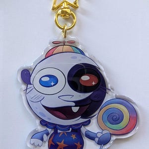 Könnte beinhalten: Ein klarer Acryl-Anhänger mit einer Cartoonfigur mit weißem Gesicht, einem halb schwarzen Auge und einem bunten Lutscher. Die Figur trägt ein blaues Outfit mit orangenen Sternen und Schuhen. Ein goldfarbener sternförmiger Verschluss ist angebracht.