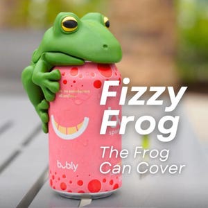 May include: A green frog-shaped can cover, with yellow eyes, is perched on a pink can of bubbly sparkling water. The can has red spots and a smiling face design. Text on the can reads "Fizzy Frog" and "The Frog Can Cover."