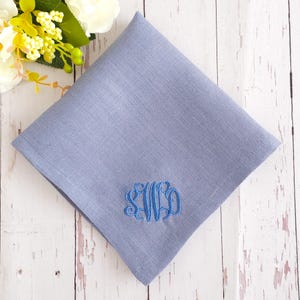 May include: A pale blue handkerchief with a matching embroidered monogram. The handkerchief is folded and placed on a light wooden surface. The monogram features the letters S, W, and D. The fabric has a subtle texture.