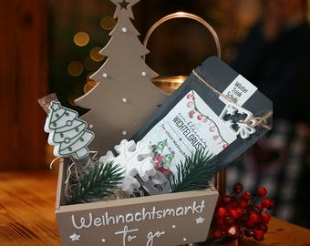 May include: A wooden box with the text "Weihnachtsmarkt To go" contains a decorative Christmas tree, a snowflake, a small bottle with a Christmas tree design, and a package with the text "Wichtelgruss". Red berries and a metal lantern are also visible.