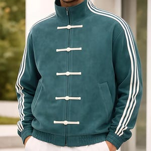 May include: Teal jacket with white stripes on the sleeves and a mandarin collar. The jacket features a zip closure and white toggle buttons. The model is wearing white trousers. The jacket has a sporty, yet classic design.