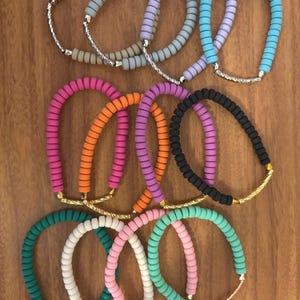 May include: A collection of colorful beaded bracelets in various shades, including pink, orange, purple, and teal. Each bracelet features a curved design with a silver or gold-toned metal accent.