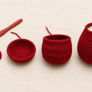 May include: A red crochet hook and four stages of a red crocheted project in progress. The pieces include a flat circular base, a shallow bowl shape, a deeper textured cup, and a finished rounded vessel with a white interior opening.