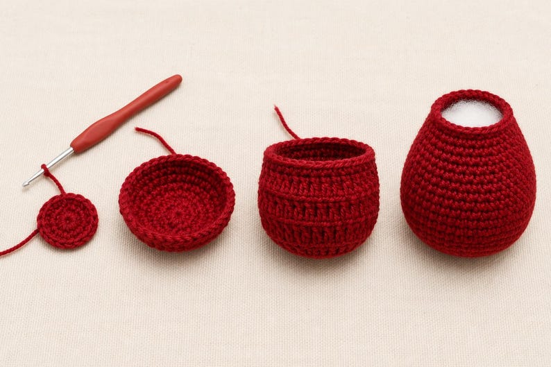 May include: A red crochet hook and four stages of a red crocheted project in progress. The pieces include a flat circular base, a shallow bowl shape, a deeper textured cup, and a finished rounded vessel with a white interior opening.