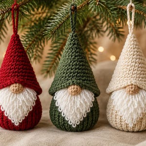 May include: Three cone-shaped crochet ornaments featuring gnomes with pointed hats in red, dark green, and cream. Each gnome has a small tan nose, a textured white yarn beard, and a matching crochet loop at the top for hanging on a tree branch.