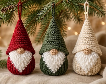 May include: Three cone-shaped crochet ornaments featuring gnomes with pointed hats in red, dark green, and cream. Each gnome has a small tan nose, a textured white yarn beard, and a matching crochet loop at the top for hanging on a tree branch.