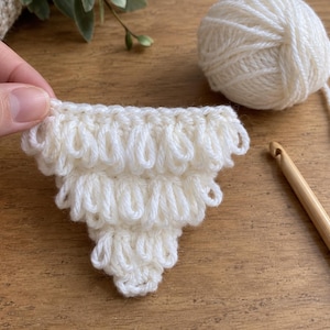 May include: A triangular crochet piece made with white yarn, featuring a textured loop stitch pattern. The piece is shown alongside a ball of matching white yarn and a wooden crochet hook, demonstrating the craft process of creating this item.