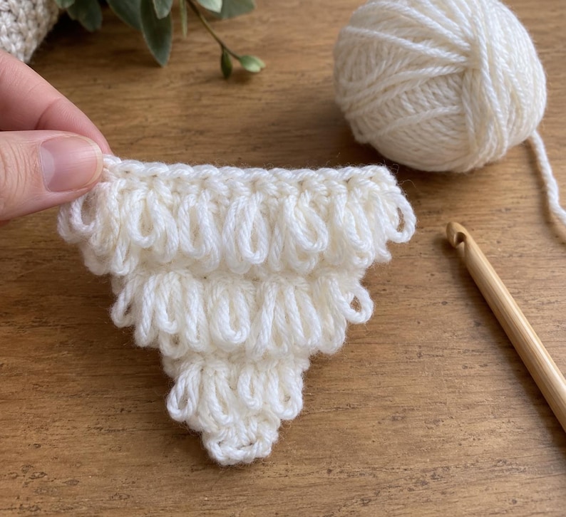 May include: A triangular crochet piece made with white yarn, featuring a textured loop stitch pattern. The piece is shown alongside a ball of matching white yarn and a wooden crochet hook, demonstrating the craft process of creating this item.