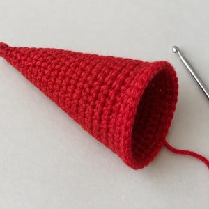 May include: A bright red cone-shaped crochet piece made with tight, uniform stitches. The hollow object tapers to a point at one end and features an open circular rim at the other. A single strand of red yarn extends from the base near a metal hook.