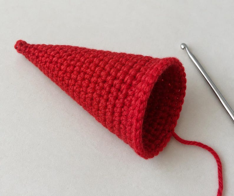 May include: A bright red cone-shaped crochet piece made with tight, uniform stitches. The hollow object tapers to a point at one end and features an open circular rim at the other. A single strand of red yarn extends from the base near a metal hook.