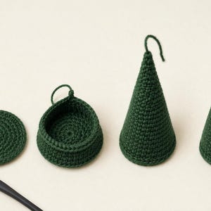 May include: A set of dark green crocheted items including a flat circular piece, a small bowl-like container, and two cone-shaped ornaments with hanging loops, accompanied by a metal crochet hook with a black handle for crafting these pieces.