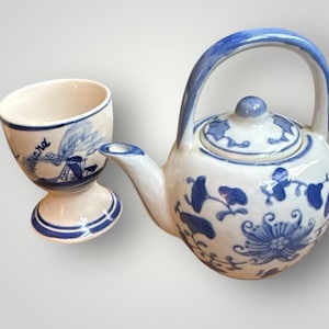 May include: A white ceramic teapot and egg cup set with blue floral designs. The teapot has a curved handle and a lid with a small knob. The egg cup features a windmill illustration and the word "Holland".
