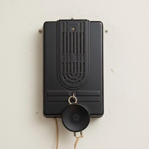 Pode incluir: Um telefone de parede preto vintage com um fone de ouvido circular e um corpo retangular. O telefone está montado em uma parede branca. A marca "EDWARDS" é visível na parte superior do telefone.