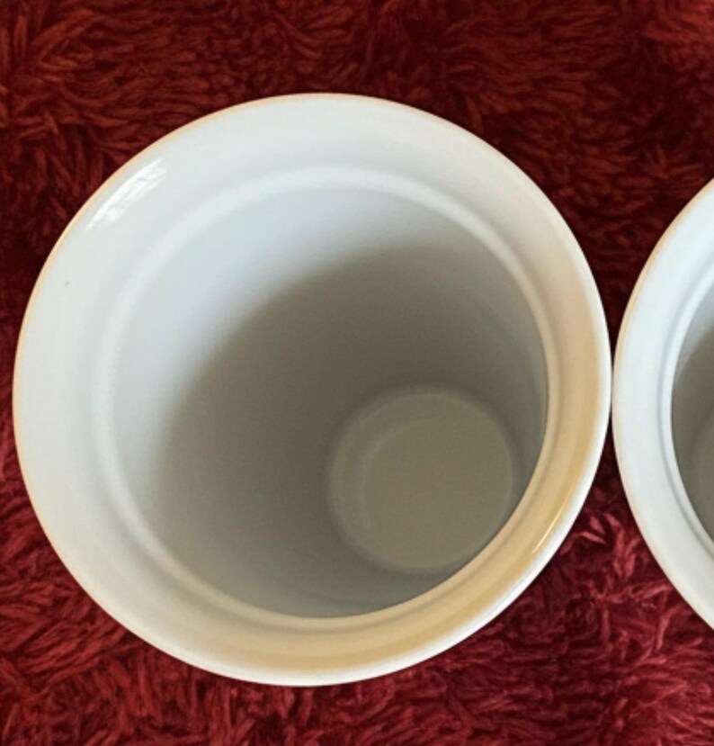 Pu&ograve; includere: Due contenitori in ceramica bianca con finitura lucida. I contenitori hanno una forma cilindrica con un'ampia apertura e una base arrotondata. L'interno &egrave; di colore grigio chiaro. I contenitori sono su uno sfondo rosso.