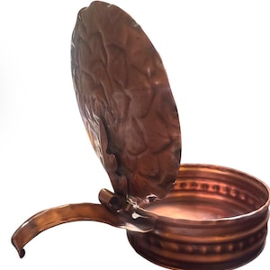 May include: A round copper container with a hinged lid that stands upright. The lid features a textured, hammered surface, while the base has a smooth interior and a decorative, embossed band around the exterior side. A curved handle extends outward.