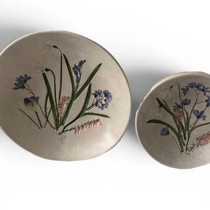 May include: Two round, off-white ceramic bowls of different sizes, each featuring a hand-painted botanical illustration of blue, white, and pink spring flowers with green stems and leaves centered on the interior surface of the dish.