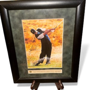 Puede incluir: Fotografía enmarcada de un golfista en acción, balanceando un palo de golf. La imagen está ambientada sobre un fondo de campo de golf con un tono cálido y dorado. El marco es negro y la fotografía incluye una firma y una cita.